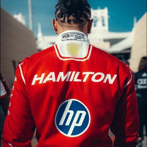 Pode incluir: Casaco de corrida vermelho com detalhes brancos e o nome "HAMILTON" em letras brancas grandes. O logotipo da HP est&aacute; no casaco, juntamente com o logotipo da Puma. O casaco tem uma gola branca com a marca FIA e Puma.