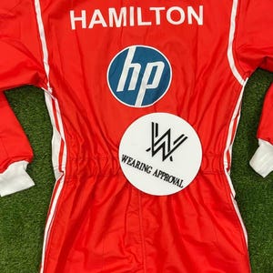 Pode incluir: Macac&atilde;o de corrida vermelho com detalhes brancos e o nome "HAMILTON" nas costas. Apresenta o logotipo da HP e um patch "WEARING APPROVAL". Gola e punhos brancos.