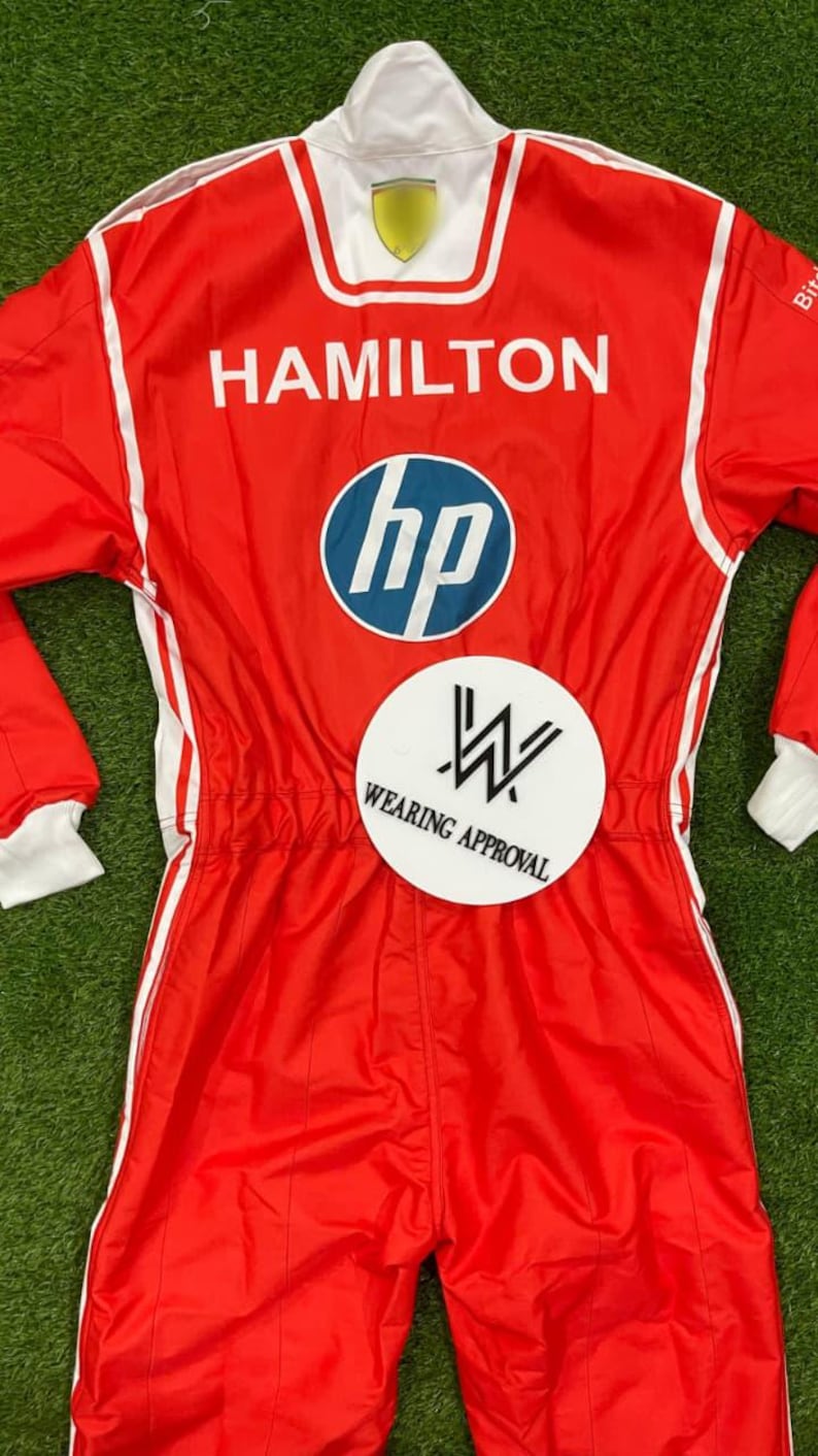 Pode incluir: Macac&atilde;o de corrida vermelho com detalhes brancos e o nome "HAMILTON" nas costas. Apresenta o logotipo da HP e um patch "WEARING APPROVAL". Gola e punhos brancos.