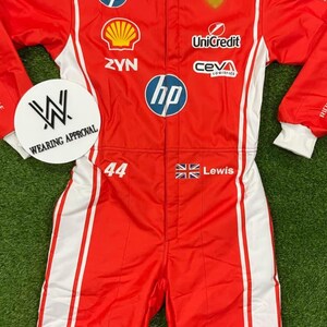 Pode incluir: Macac&atilde;o de corrida vermelho e branco com logotipos de patrocinadores, incluindo HP, Shell e IBM. O macac&atilde;o apresenta o n&uacute;mero 44, o nome "Lewis" e uma bandeira brit&acirc;nica. Listras brancas nas laterais. Em uma superf&iacute;cie verde.