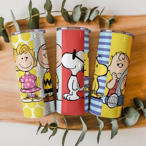 Puede incluir: Tres vasos de acero inoxidable con personajes de Peanuts. Un vaso tiene a Sally Brown, otro a Charlie Brown y el tercero a Snoopy. Los vasos tienen bloques de color amarillo, rojo y azul con fondos estampados.