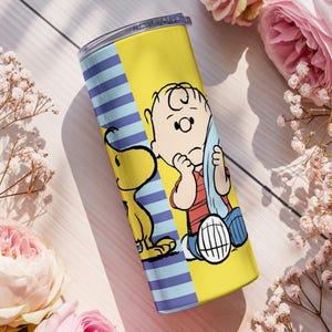 Puede incluir: Un vaso amarillo y azul con Charlie Brown y Snoopy de la tira c&oacute;mica Peanuts. El vaso tiene un fondo amarillo con una secci&oacute;n de rayas azules. Los personajes est&aacute;n a todo color, con Charlie Brown con aspecto triste y Snoopy a su lado.