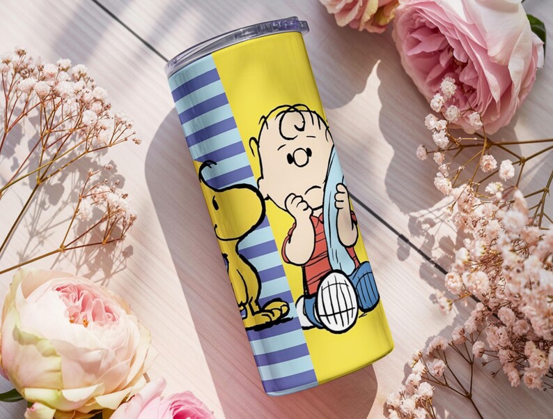 Puede incluir: Un vaso amarillo y azul con Charlie Brown y Snoopy de la tira c&oacute;mica Peanuts. El vaso tiene un fondo amarillo con una secci&oacute;n de rayas azules. Los personajes est&aacute;n a todo color, con Charlie Brown con aspecto triste y Snoopy a su lado.