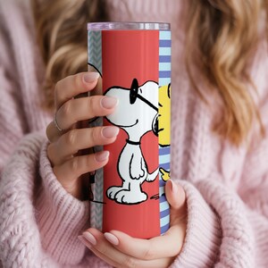 Puede incluir: Un vaso de acero inoxidable con un dise&ntilde;o de dibujos animados de Snoopy. El vaso tiene un fondo rojo con Snoopy con gafas. El dise&ntilde;o tambi&eacute;n incluye una secci&oacute;n de rayas azules y blancas. El vaso lo sostiene una persona que lleva un su&eacute;ter rosa.