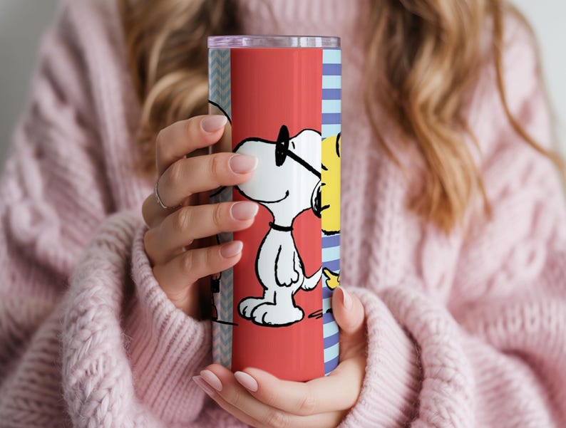 Puede incluir: Un vaso de acero inoxidable con un dise&ntilde;o de dibujos animados de Snoopy. El vaso tiene un fondo rojo con Snoopy con gafas. El dise&ntilde;o tambi&eacute;n incluye una secci&oacute;n de rayas azules y blancas. El vaso lo sostiene una persona que lleva un su&eacute;ter rosa.