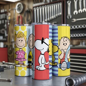 Puede incluir: Cuatro vasos de acero inoxidable con personajes de Peanuts. Los vasos tienen dise&ntilde;os coloridos, incluyendo lunares, rayas e ilustraciones de personajes. El fondo muestra un taller con herramientas.
