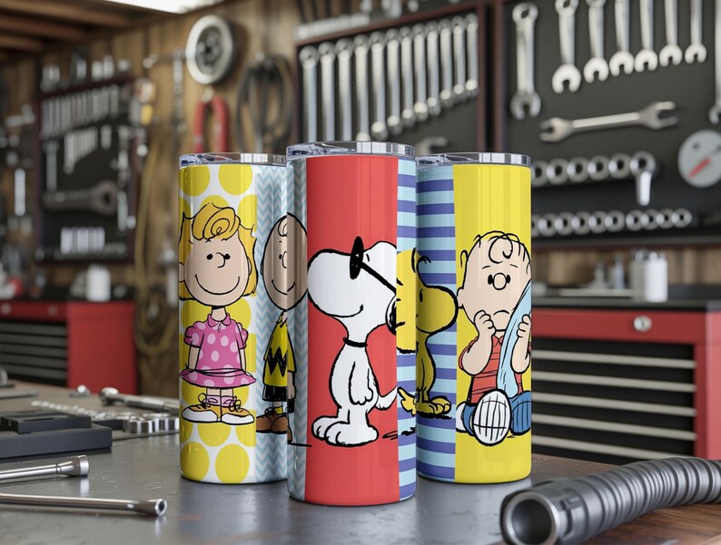 Puede incluir: Cuatro vasos de acero inoxidable con personajes de Peanuts. Los vasos tienen dise&ntilde;os coloridos, incluyendo lunares, rayas e ilustraciones de personajes. El fondo muestra un taller con herramientas.