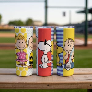 Puede incluir: Cuatro vasos de acero inoxidable con personajes de Peanuts. Cada vaso tiene un dise&ntilde;o diferente: amarillo con lunares, blanco con zigzags, rojo con Snoopy y amarillo con Charlie Brown. Los vasos est&aacute;n sobre una superficie de madera.