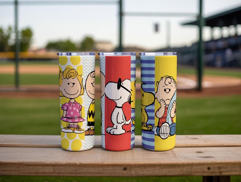 Puede incluir: Cuatro vasos de acero inoxidable con personajes de Peanuts. Cada vaso tiene un dise&ntilde;o diferente: amarillo con lunares, blanco con zigzags, rojo con Snoopy y amarillo con Charlie Brown. Los vasos est&aacute;n sobre una superficie de madera.