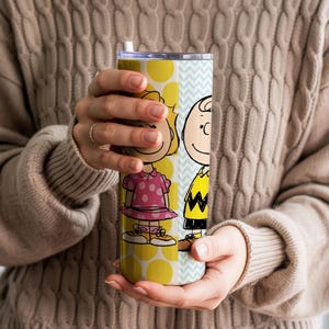 Puede incluir: Un vaso amarillo y azul claro con los personajes de dibujos animados Sally Brown y Charlie Brown. El vaso tiene una pajita blanca y un estampado de lunares amarillos. Los personajes visten sus trajes caracter&iacute;sticos.
