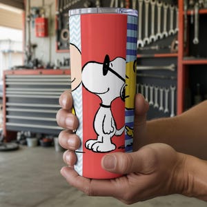 Puede incluir: Un vaso rojo y blanco con Snoopy con gafas de sol, con Charlie Brown y Woodstock en los lados. El vaso tiene una tapa plateada y lo sostiene una persona. El fondo es un garaje.
