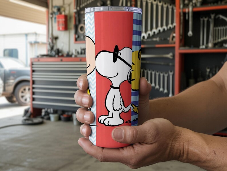 Puede incluir: Un vaso rojo y blanco con Snoopy con gafas de sol, con Charlie Brown y Woodstock en los lados. El vaso tiene una tapa plateada y lo sostiene una persona. El fondo es un garaje.