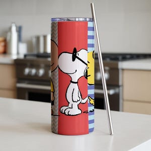 Puede incluir: Un vaso de acero inoxidable con un dise&ntilde;o de dibujos animados de Snoopy y Woodstock sobre un fondo rojo. El vaso tiene una pajita plateada y est&aacute; colocado sobre una encimera blanca. El dise&ntilde;o incluye un patr&oacute;n de chevr&oacute;n y rayas azules.