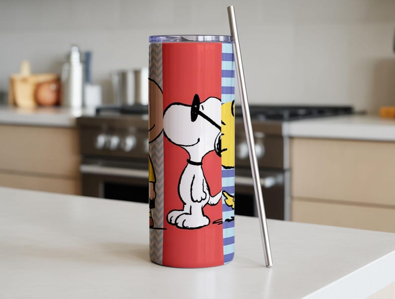Puede incluir: Un vaso de acero inoxidable con un dise&ntilde;o de dibujos animados de Snoopy y Woodstock sobre un fondo rojo. El vaso tiene una pajita plateada y est&aacute; colocado sobre una encimera blanca. El dise&ntilde;o incluye un patr&oacute;n de chevr&oacute;n y rayas azules.