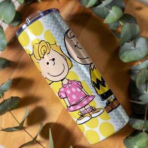 Puede incluir: Un vaso de acero inoxidable con los personajes de dibujos animados Sally y Charlie Brown. El vaso tiene un patr&oacute;n de lunares amarillos y galones azules. Sally lleva un vestido rosa y Charlie Brown una camisa amarilla con un dise&ntilde;o en zigzag.