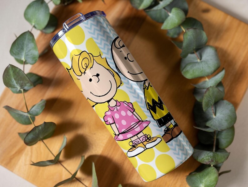 Puede incluir: Un vaso de acero inoxidable con los personajes de dibujos animados Sally y Charlie Brown. El vaso tiene un patr&oacute;n de lunares amarillos y galones azules. Sally lleva un vestido rosa y Charlie Brown una camisa amarilla con un dise&ntilde;o en zigzag.