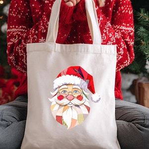 Peut inclure: Un sac fourre-tout crème avec un motif de visage de Père Noël coloré. Le visage du Père Noël a un chapeau rouge, des lunettes et une barbe blanche. Le sac est tenu par une personne portant un pull rouge.