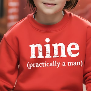 Peut inclure: Sweat-shirt rouge avec le mot "nine" en grandes lettres blanches usées, suivi de "(practically a man)". Le sweat-shirt est de couleur rouge uni.