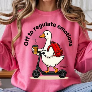 May include: Pink sweatshirt featuring a cartoon goose on a scooter, carrying a coffee and a red backpack. The text above the goose reads "Off to regulate emotions".