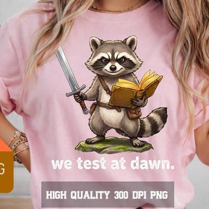 Peut inclure: T-shirt rose avec un raton laveur de dessin animé tenant une épée et un livre. Le raton laveur est sur un rocher vert. Le texte "we test at dawn" est en dessous. L'image comprend également le texte "HIGH QUALITY 300 DPI PNG."