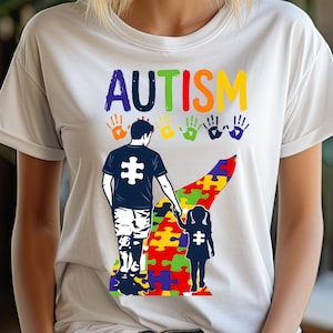 May include: White t-shirt featuring the word "AUTISM" in colourful letters above handprints and a graphic of a parent and child holding hands. The parent and child wear shirts with puzzle piece graphics.