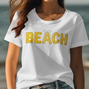May include: White t-shirt with the word "BEACH" in yellow, textured letters. The letters are outlined in a darker color, and the "A" is decorated with a bow. The shirt is worn by a person.