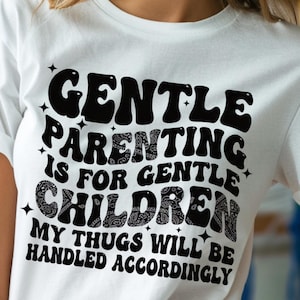 Peut inclure: T-shirt blanc avec l'inscription noire "GENTLE PARENTING IS FOR GENTLE CHILDREN. MY THUGS WILL BE HANDLED ACCORDINGLY" dans une police rétro. Le design comprend des étoiles.