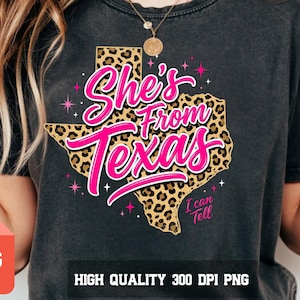 Puede incluir: Camiseta gris oscuro con un gráfico rosa y blanco que dice "She's From Texas" con un contorno de Texas con estampado de leopardo. También se incluye el texto "I can tell".