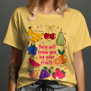 Peut inclure: T-shirt jaune avec un motif de fruits colorés. Le design comprend des bananes, des cerises, une poire, des myrtilles, de la pastèque, une orange, une pêche, des raisins et des fraises. Le texte dit "they will know you by your fruits."