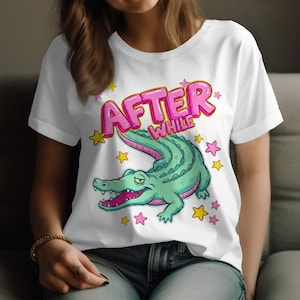 Peut inclure: T-shirt blanc avec un dessin de crocodile de dessin animé et le texte "AFTER WHILE". Le crocodile est vert avec des détails roses. Des étoiles roses et jaunes entourent le dessin.