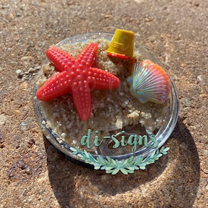 May include: A clear glass tumbler with a red starfish, a yellow bucket, and a iridescent seashell on a bed of white sand. The tumbler has the text "di-signs" on the bottom.