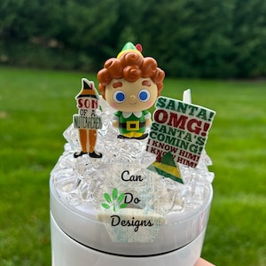 May include: A white tumbler with a pink handle, adorned with Christmas-themed decorations. The decorations include a cartoon elf figure and signs that read "SON OF A NUTCRACKER" and "SANTA! OMG! SANTA'S COMING! I KNOW HIM!" The tumbler has the text "Can Do Designs".