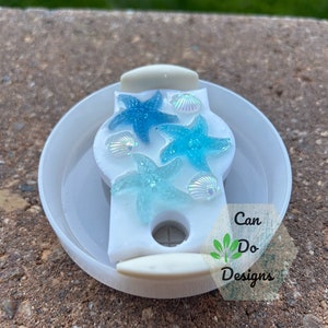 May include: White jar lid with a blue and green starfish design and iridescent seashells. The lid has a hole in the center and the text "Can Do Designs" is printed on the side.