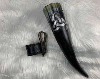 May include: A black and white drinking horn with a silver triquetra design. The horn is accompanied by a black leather strap with a silver hammer design.