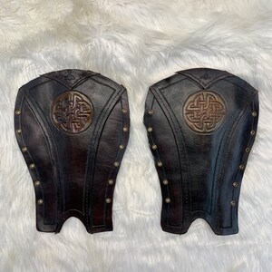 May include: A pair of brown leather leg guards with a Celtic knot design. The guards are secured with brass rivets.