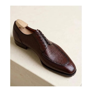 May include: A dark brown leather oxford shoe with a distinct pebbled texture, featuring a stitched apron toe, a closed lacing system with thin dark laces, and a light-colored wooden shoe tree inserted into the heel area for shape retention.