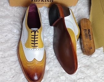 May include: A pair of wingtip oxford shoes featuring a multi-coloured design in white, yellow, and brown with decorative brogue detailing. The shoes have dark brown laces and a smooth finish. A wooden shoe brush and a cardboard box are included.