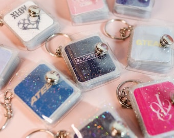 May include: A collection of square mini photo album keychains with clear plastic covers and silver metal snap closures. Each keychain features a unique colored insert with various logos and text including BLACKPINK, ATEEZ, and other graphic designs.