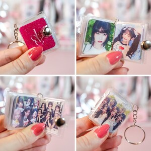May include: Clear acrylic keychain with a silver metal ring. The keychain features a small, square photo album with a pink cover and a silver clasp. The album contains photos of people. The keychain is held by a person with red nail polish.