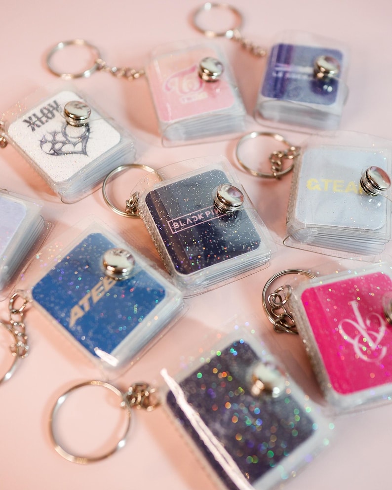 May include: A collection of small, glittery keychains with various designs and text, including the words "BLACKPINK" and "ATEEZ". Each keychain features a clear plastic cover and a silver-colored metal ring.