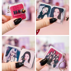 May include: A clear, glittery keychain featuring a small, open book design. The cover is red with the text "BABYMONSTER" and a button closure. Inside are photos of a group of women. The keychain is attached to a silver ring.