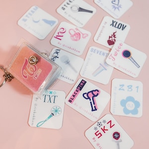 May include: A collection of white, rectangular stickers featuring various K-pop group logos and light stick illustrations. A clear, glittery keychain with a pink TWICE logo is also present. The stickers are scattered on a pink surface.
