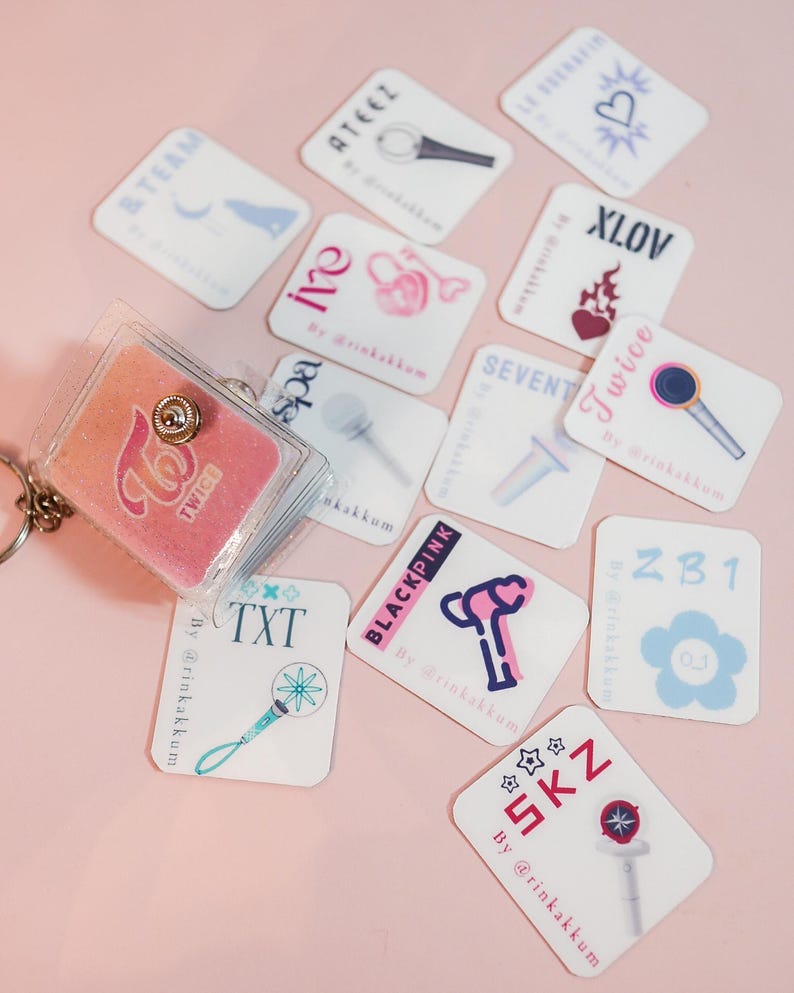 May include: A collection of white, rectangular stickers featuring various K-pop group logos and light stick illustrations. A clear, glittery keychain with a pink TWICE logo is also present. The stickers are scattered on a pink surface.