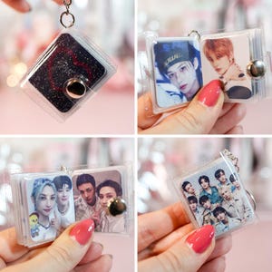 May include: A clear, square keychain with a silver metal clasp. The keychain holds a small photo album with multiple images of a K-pop group. The album is held open by a clear plastic cover.
