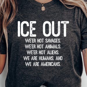Könnte beinhalten: Dunkelgraues Unisex-T-Shirt mit dem weißen Schriftzug "ICE OUT" in großen Buchstaben. Darunter steht in kleinerer weißer Schrift: "WE'ER NOT SAVAGES. WE'ER NOT ANIMALS. WE'ER NOT ALIENS. WE ARE HUMANS, AND WE ARE AMERICANS."