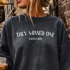 May include: A charcoal gray crewneck sweatshirt featuring white text that reads "THEY MISSED ONE" above the smaller text "SALEM, 1692". The long-sleeved garment has a relaxed fit and is paired with light blue denim shorts with a frayed hem.
