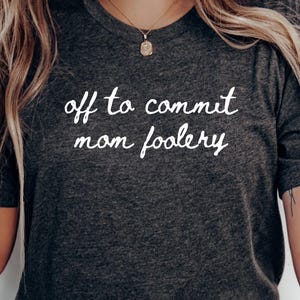 Könnte beinhalten: Ein dunkel anthrazitfarbenes Kurzarm-T-Shirt mit dem weißen Schriftzug "off to commit mom foolery" in einer lässigen Schreibschrift auf der Brust. Das T-Shirt hat einen klassischen Rundhalsausschnitt und eine melierte Stoffstruktur.