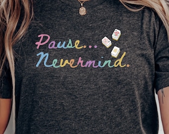 May include: A dark heather gray short-sleeve t-shirt featuring the text "Pause... Nevermind." in a multi-colored pastel font. Three small mahjong tiles are positioned above the word "Nevermind" on the front of the garment.