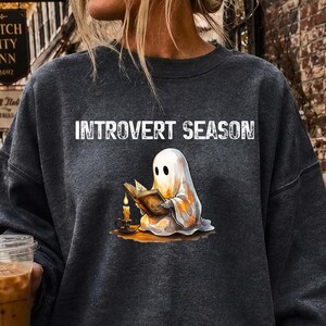 Puede incluir: Una sudadera de cuello redondo en color gris carbón oscuro con un gráfico de un fantasma blanco leyendo un libro a la luz de una vela. El texto INTROVERT SEASON está impreso en blanco sobre la ilustración del fantasma. Tiene mangas largas.