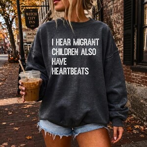 May include: A dark charcoal gray crewneck sweatshirt featuring the white text I HEAR MIGRANT CHILDREN ALSO HAVE HEARTBEATS printed on the front. The garment has long sleeves and a relaxed fit, paired with light blue denim shorts.